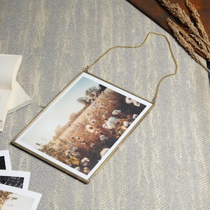 以下が含まれることがあります： 上部にチェーンがぶら下がっている金色の金属製のフォトフレーム。フレームには、白い綿の植物の畑の写真が入っています。