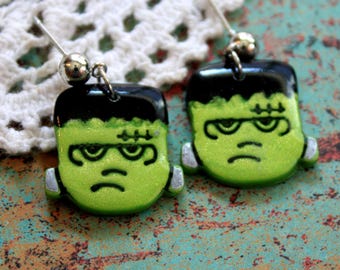 May include: A pair of handmade Halloween earrings featuring a green Frankenstein monster design. The earrings have a black top, silver accents, and a silver ball at the top. The background is a textured, multi-colored surface.