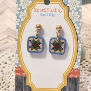 May include: A pair of square earrings with a blue border and a geometric pattern in shades of green, brown, and red. The earrings are suspended from gold-colored ball studs. The product is displayed on a decorative card with the text "Knot2Shabby Bags & Brags".