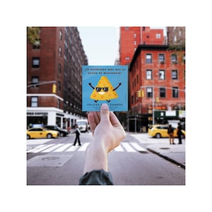 May include: A square blue card held up in front of a city street. The card features a cartoon slice of cheese wearing sunglasses and the text "¡TE QUEREMOS MAS QUE EL QUESO DE WISCONSIN!" and "FELICES SIEMPRE".