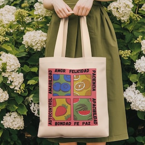 May include: A cream-colored tote bag with a colorful design featuring the words "Amor, Felicidad, Paciencia, Apacibilidad, Bondad, Fe, Paz, and Autocontrol" and illustrations of blueberries, oranges, strawberries, and watermelon. The bag has cream-colored handles.