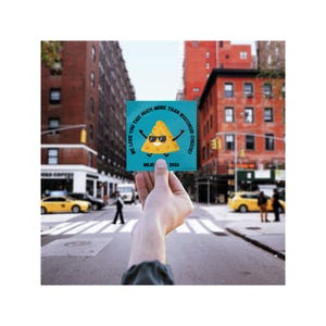 May include: A square, teal-colored sticker held up in front of a city street. The sticker features a cartoon nacho chip wearing sunglasses and the text "WE LOVE YOU THIS MUCH MORE THAN WISCONSIN CHEESE!" with "MILW. 2026" below.