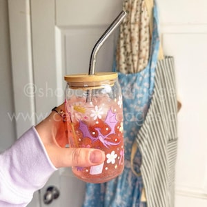 May include: A clear glass tumbler with a bamboo lid and a bent metal straw, filled with a pink liquid. The glass features a printed design of a purple dragon, a gold sword, a gold crown, white flowers, and small stars scattered across the surface.