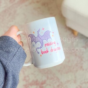 May include: A white ceramic mug featuring a purple dragon illustration and the text "raising book dragons" in pink and red lettering. The design includes small orange and pink star-shaped accents surrounding the dragon and the printed text.