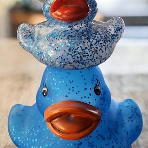 May include: Two blue rubber duck figurines, one stacked on top of the other. The bottom duck is a solid blue, while the top duck is covered in blue glitter and wears a small, light-coloured hat. Both have orange beaks.