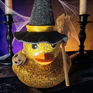 May include: A yellow glittery rubber duck wearing a black witch hat with a yellow ribbon band. The duck features a bright orange beak, black eyes, a small carved pumpkin figure on its side, and a miniature wooden broomstick held upright.