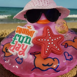 May include: A pink rubber duck wearing a light pink woven hat and black sunglasses. It features a white sign with the orange, green, and pink text "SUNDAY fun day," a red starfish, and a small clear glass filled with a yellow liquid.