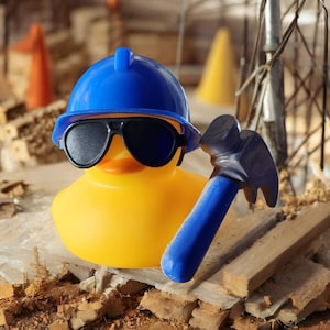 Pode incluir: Um pato de borracha amarelo usando um capacete de construção azul e óculos de sol pretos, segurando um martelo azul. O pato está em um cenário de construção com tijolos e cones.