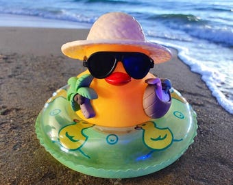 May include: A bright yellow rubber duck wearing a tan straw hat and black sunglasses. The duck holds a small green palm tree and a brown coconut drink while sitting inside a translucent green inflatable swim ring decorated with yellow duck prints.