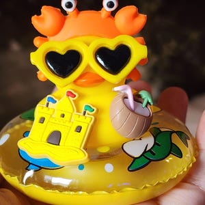 May include: A yellow rubber duck wearing an orange crab hat and yellow heart-shaped sunglasses. The duck sits in a yellow inflatable ring decorated with a yellow sandcastle, a brown coconut drink with a straw, and green palm leaf graphics.