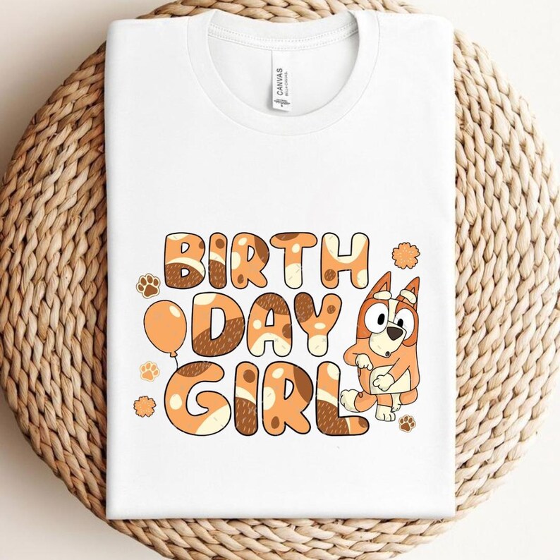 May include: White t-shirt with the words "BIRTH DAY GIRL" in brown and orange bubble letters, featuring a cartoon dog character. The shirt has a round neck and short sleeves. A paw print and a balloon are also included in the design.