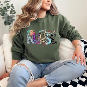 May include: An olive green sweatshirt featuring a colourful "NURSE" graphic. The design incorporates floral, leopard print, and medical symbols such as a syringe and stethoscope. The letters are in diverse patterns and colours.