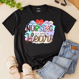 May include: Black t-shirt featuring the text "NURSING is a work of Heart" in a colourful design. The graphic includes a heartbeat line and two red hearts. The shirt is shown with blue jeans and sandals.