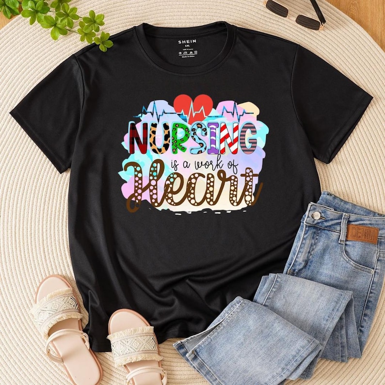 May include: Black t-shirt featuring the text "NURSING is a work of Heart" in a colourful design. The graphic includes a heartbeat line and two red hearts. The shirt is shown with blue jeans and sandals.