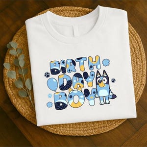 May include: White t-shirt with the words "BIRTH DAY BOY" in blue, yellow, and white bubble letters. A cartoon dog character is on the right. Paw prints and a blue balloon are also featured in the design.