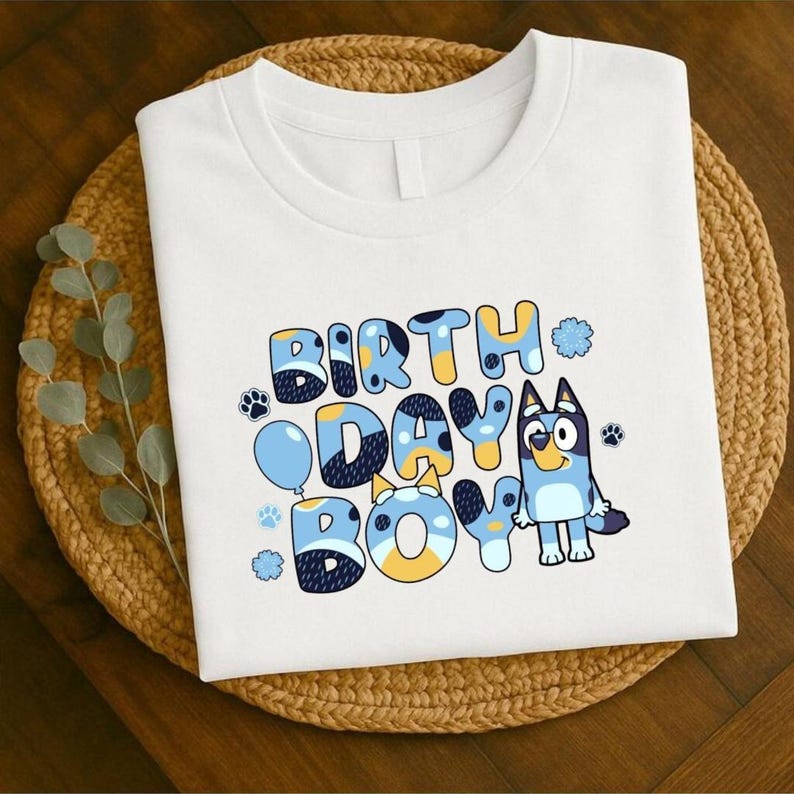 May include: White t-shirt with the words "BIRTH DAY BOY" in blue, yellow, and white bubble letters. A cartoon dog character is on the right. Paw prints and a blue balloon are also featured in the design.