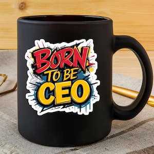 May include: A black ceramic mug with a large handle. The mug features the text "BORN TO BE CEO" in a colorful, stylized design with red, yellow, and blue accents. The mug is sitting on a patterned surface.