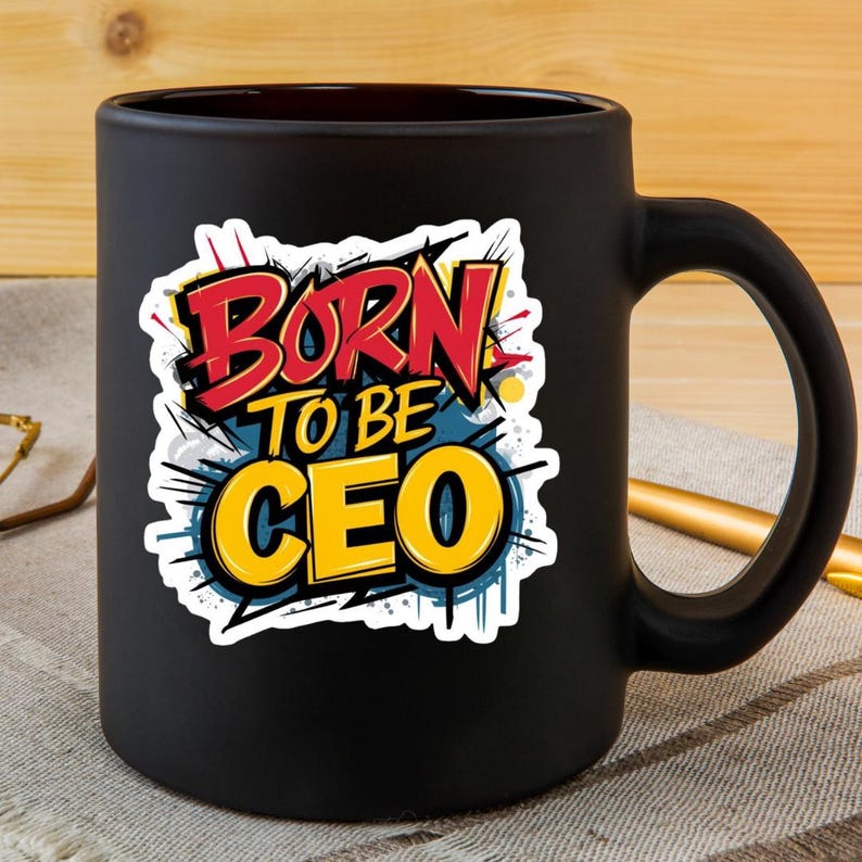 May include: A black ceramic mug with a large handle. The mug features the text "BORN TO BE CEO" in a colorful, stylized design with red, yellow, and blue accents. The mug is sitting on a patterned surface.