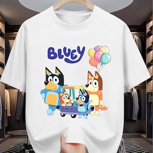 May include: White t-shirt featuring the word "Bluey" in blue script above a cartoon image of Bluey and her family. The cartoon characters are in a car and holding balloons.