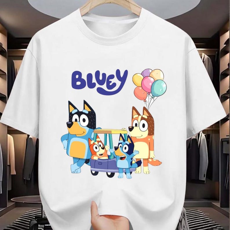 May include: White t-shirt featuring the word "Bluey" in blue script above a cartoon image of Bluey and her family. The cartoon characters are in a car and holding balloons.