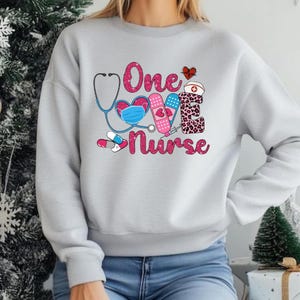 May include: A pale grey sweatshirt featuring the text "One Love Nurse" in pink, accompanied by medical-themed illustrations. The design incorporates a stethoscope, heart, face mask, bandage, syringe, pills, and a nurse's hat.