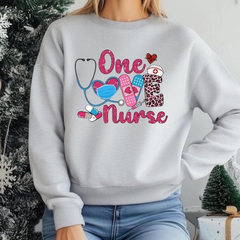 May include: A pale grey sweatshirt featuring the text "One Love Nurse" in pink, accompanied by medical-themed illustrations. The design incorporates a stethoscope, heart, face mask, bandage, syringe, pills, and a nurse's hat.
