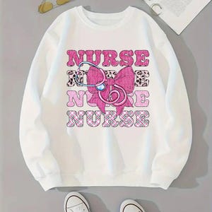 May include: White sweatshirt featuring the word "NURSE" in pink block letters, a pink bow, and a stethoscope graphic. The letters have a leopard print and plaid pattern. The sweatshirt is a casual style.