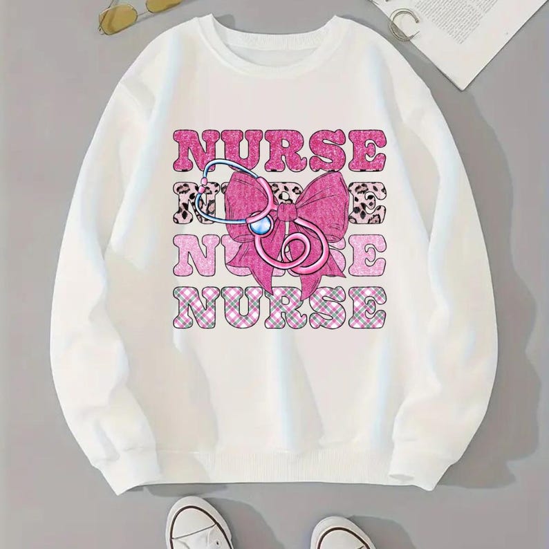 May include: White sweatshirt featuring the word "NURSE" in pink block letters, a pink bow, and a stethoscope graphic. The letters have a leopard print and plaid pattern. The sweatshirt is a casual style.