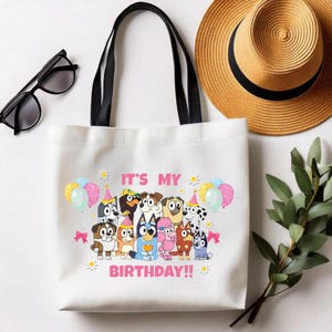 May include: White tote bag with black handles featuring a colorful graphic of cartoon dogs and the text "IT'S MY BIRTHDAY!!" Balloons and bows decorate the design. A straw hat and sunglasses are in the background.