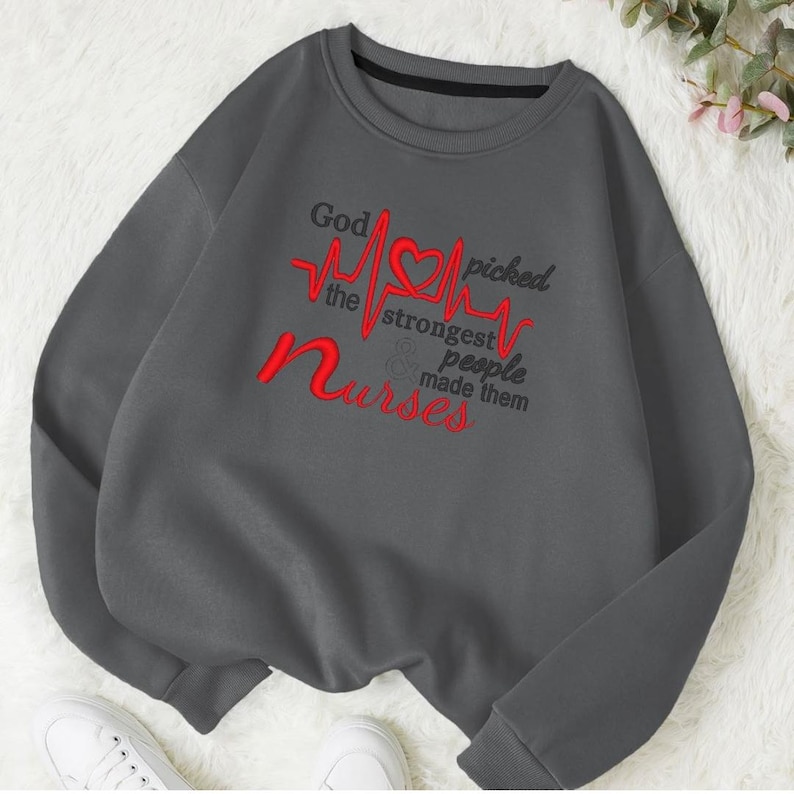 May include: A charcoal grey sweatshirt featuring the embroidered phrase "God picked the strongest people & made them nurses" in red and black. The design includes a red heart and a heartbeat line. It has a crew neck and long sleeves.