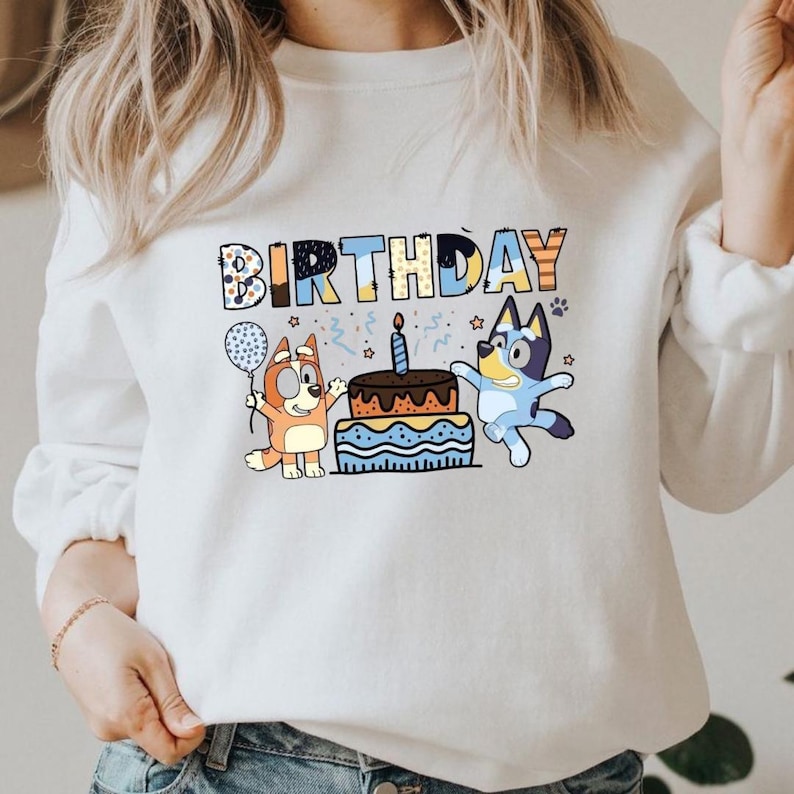 May include: White sweatshirt featuring the word "BIRTHDAY" in colorful block letters. The design includes cartoon characters, a birthday cake with a lit candle, and a balloon. The characters are in shades of orange and blue.
