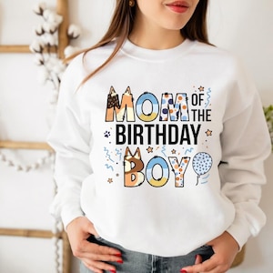 May include: White sweatshirt with the text "MOM OF THE BIRTHDAY BOY" in colorful, patterned letters. The design includes stars, paw prints, and a blue balloon. A festive and celebratory garment.