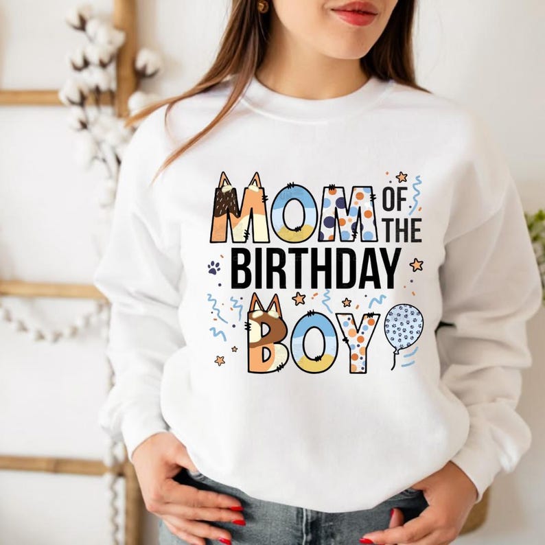 May include: White sweatshirt with the text "MOM OF THE BIRTHDAY BOY" in colorful, patterned letters. The design includes stars, paw prints, and a blue balloon. A festive and celebratory garment.