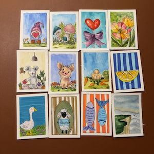 May include: A collection of watercolor paintings on rectangular cards. The paintings depict various subjects, including a gnome holding a heart, mushrooms, a heart-shaped balloon, tulips, a mouse with plants, a pig, carrots, a lemon slice, a goose, a sheep with the word "found", fish, and a cliffside.