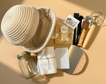 May include: A sourdough bread making kit including a round rattan proofing basket with a fabric liner, a glass jar, a white cloth, a plastic scraper, a metal dough scraper, a silicone brush, a metal whisk with a wooden handle, and a bread lame.