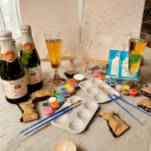 Può includere: Una natura morta per un evento di pittura e sorseggio. L'immagine presenta due tele su piccoli cavalletti, tavolozze di vernici con vari colori, pennelli e bottiglie di sidro frizzante. Sono presenti anche due bicchieri di sidro e una candela accesa.