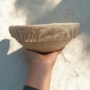 Puede incluir: Una cesta redonda de madera clara para fermentar pan con un forro de lino beige. La cesta se sostiene en una mano, mostrando su dise&ntilde;o y textura. La cesta est&aacute; dise&ntilde;ada para fermentar masa.