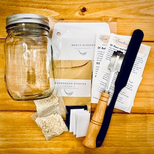 Puede incluir: Un kit de masa madre con un frasco de vidrio transparente, un sobre de papel marrón, una rasqueta de banco negra y plateada, una rasqueta de masa con mango de madera y dos pequeñas bolsas de masa madre. El kit también incluye un pequeño trozo de tela negra y cuatro etiquetas blancas.