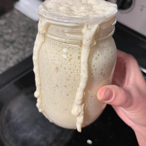 Puede incluir: Un tarro de vidrio transparente lleno de una sustancia burbujeante de color blanquecino, que se desborda y gotea por los lados. El tarro est&aacute; sostenido por una mano, lo que sugiere un alimento casero, posiblemente masa madre.