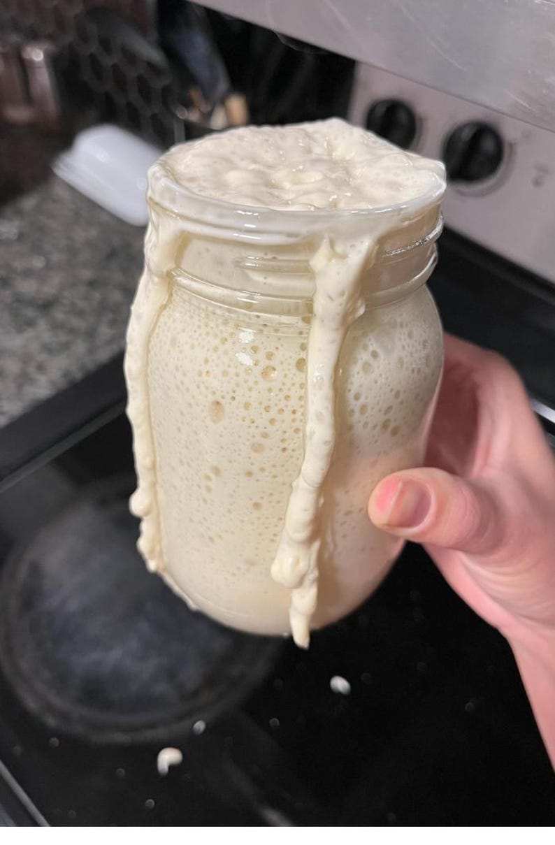 Puede incluir: Un tarro de vidrio transparente lleno de una sustancia burbujeante de color blanquecino, que se desborda y gotea por los lados. El tarro est&aacute; sostenido por una mano, lo que sugiere un alimento casero, posiblemente masa madre.