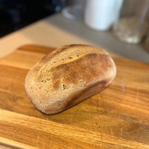 May include: A loaf of freshly baked bread with a golden-brown crust rests on a wooden cutting board. The bread has a rustic appearance, with a slightly uneven surface. The cutting board is made of light-colored wood.