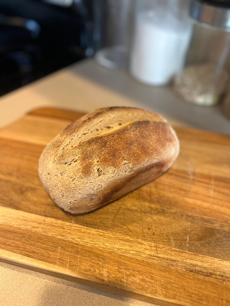 May include: A loaf of freshly baked bread with a golden-brown crust rests on a wooden cutting board. The bread has a rustic appearance, with a slightly uneven surface. The cutting board is made of light-colored wood.