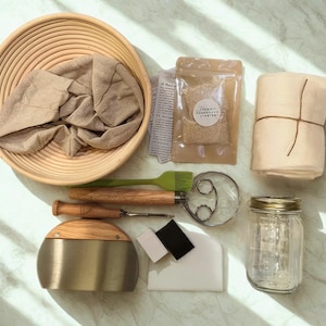 以下が含まれることがあります： 発酵かご、リネンライナー、有機スターター、瓶、生地スクレーパー、その他のツールを備えた、サワードウパン作りのキット。このキットには、木製の柄付き生地泡立て器、ベンチスクレーパー、シリコンブラシが含まれています。アイテムは明るい色の表面に配置されています。