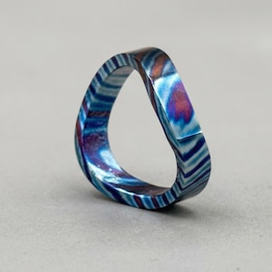 Könnte beinhalten: Ein einzigartiger Ring mit einem markanten, abstrakten Design. Der Ring weist eine Mischung aus blauen, weißen und violetten Farbtönen auf, die einen Marmoreffekt erzeugen. Die Form des Rings ist eine durchgehende Schleife mit glatter, polierter Oberfläche. Ein modernes Schmuckstück.