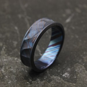 Könnte beinhalten: Ein strukturierter Ring mit einem einzigartigen Design. Der Ring hat eine facettierte Außenfläche mit Blau- und Grautönen und einen farbenfrohen, gestreiften Innenraum. Das Band ist dunkel und bildet einen auffälligen Kontrast zu den Innenfarben.