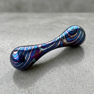 May include: A small, metallic, dumbbell-shaped object with a swirling pattern of blue, purple, and silver. The object has rounded ends connected by a narrower, curved center. The surface reflects light, creating a vibrant, iridescent effect.
