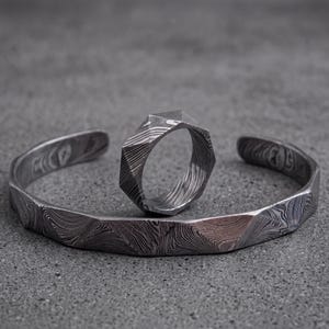 Op de afbeelding: Een zilverkleurige manchetarmband en ring set. De armband is open met een geometrisch ontwerp. De ring heeft een vergelijkbare geometrische vorm. Beide stukken hebben een patroon, een oppervlak met textuur en worden weergegeven op een grijze ondergrond.