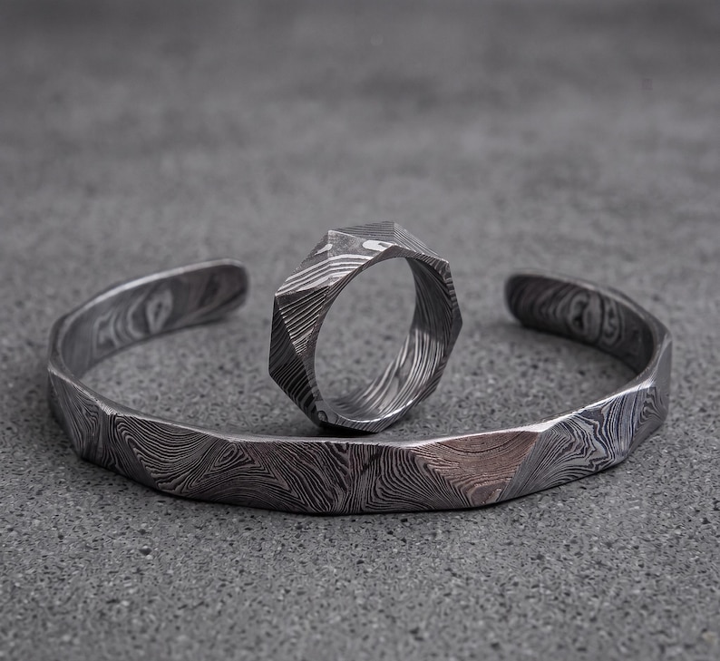 Op de afbeelding: Een zilverkleurige manchetarmband en ring set. De armband is open met een geometrisch ontwerp. De ring heeft een vergelijkbare geometrische vorm. Beide stukken hebben een patroon, een oppervlak met textuur en worden weergegeven op een grijze ondergrond.