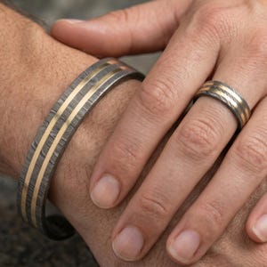 Op de afbeelding: Een armband- en ringenset van damaststaal. De sieraden hebben een donker, patroonstaal met drie parallelle goudkleurige strepen. De ring zit aan een vinger en de armband om een pols.