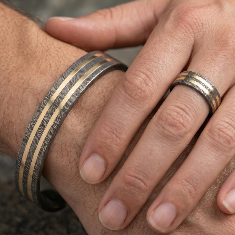 Op de afbeelding: Een armband- en ringenset van damaststaal. De sieraden hebben een donker, patroonstaal met drie parallelle goudkleurige strepen. De ring zit aan een vinger en de armband om een pols.